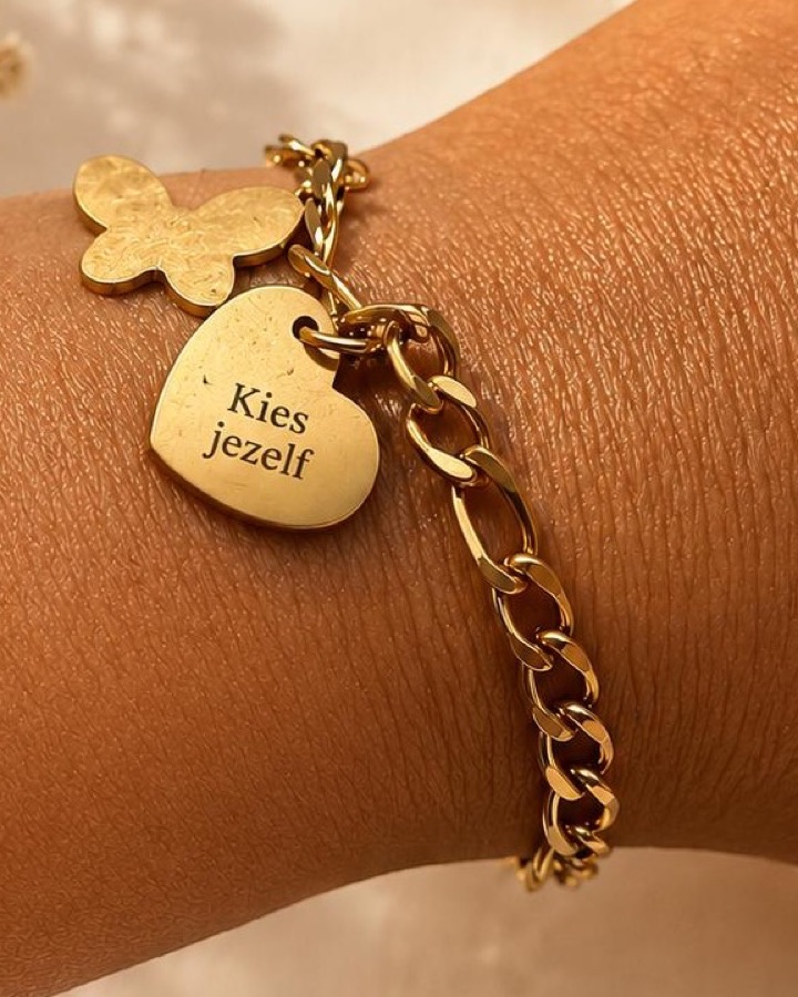 Goudkleurig schakelarmbandje om een pols, met een vlinderbedel en een hartvormige bedel met de gravure “Kies jezelf”.